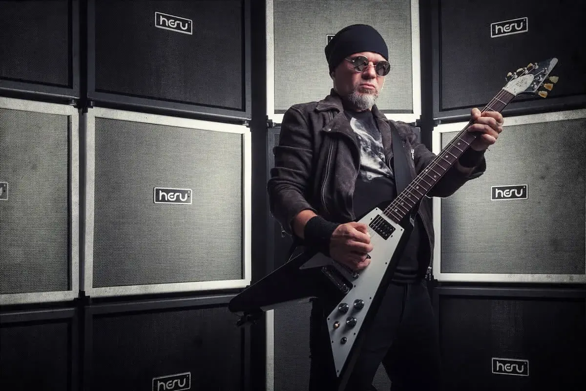 Mężczyzna w czapce i okularach gra na gitarze elektrycznej przed ścianą wzmacniaczy. Wygląda jakby był gotowy na koncert dla fanów popcorn acid drinkers.