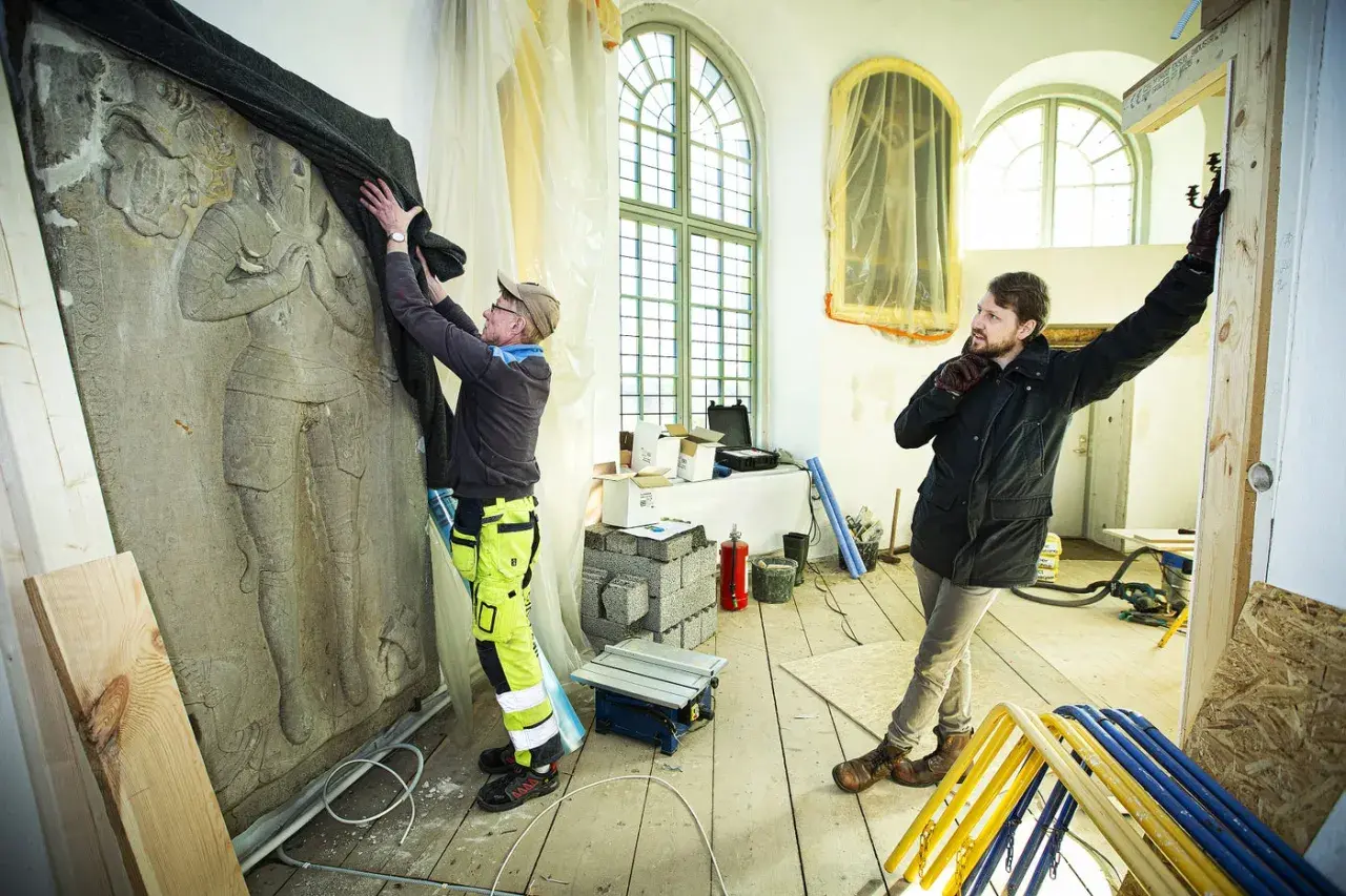 Två män arbetar med en stor stenrelief i en halvrund del av kyrka under renovering.