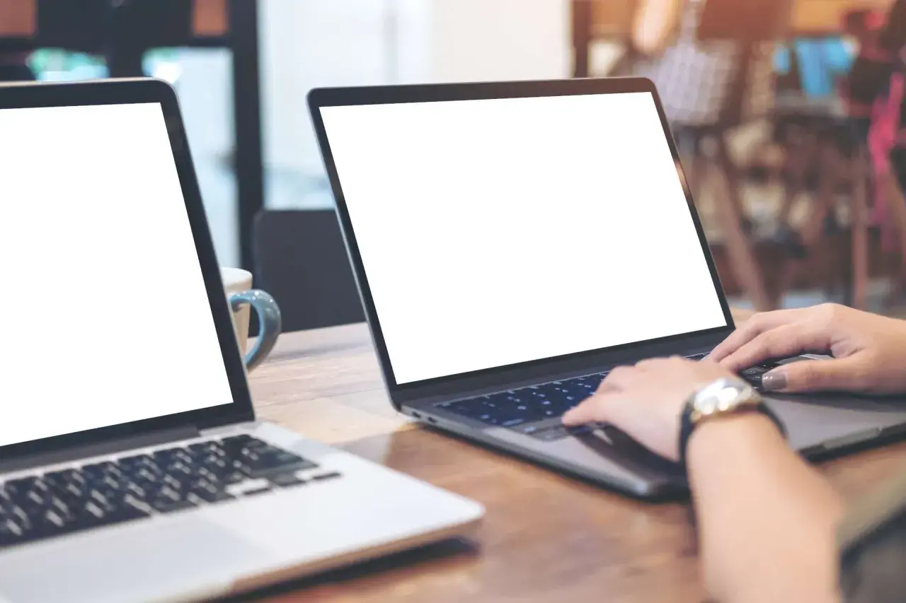 Zwei Laptops auf einem Holztisch. Eine Person nutzt einen Laptop als Monitor, während die Hände auf der Tastatur liegen.