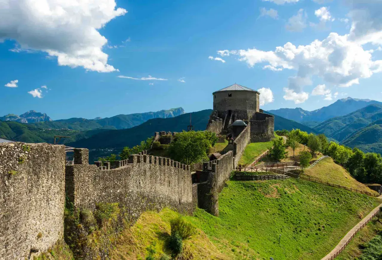 Roccia imponente e mura antiche del **San Romano in Garfagnana** si stagliano su un paesaggio verdeggiante, sotto un cielo azzurro con nuvole bianche.