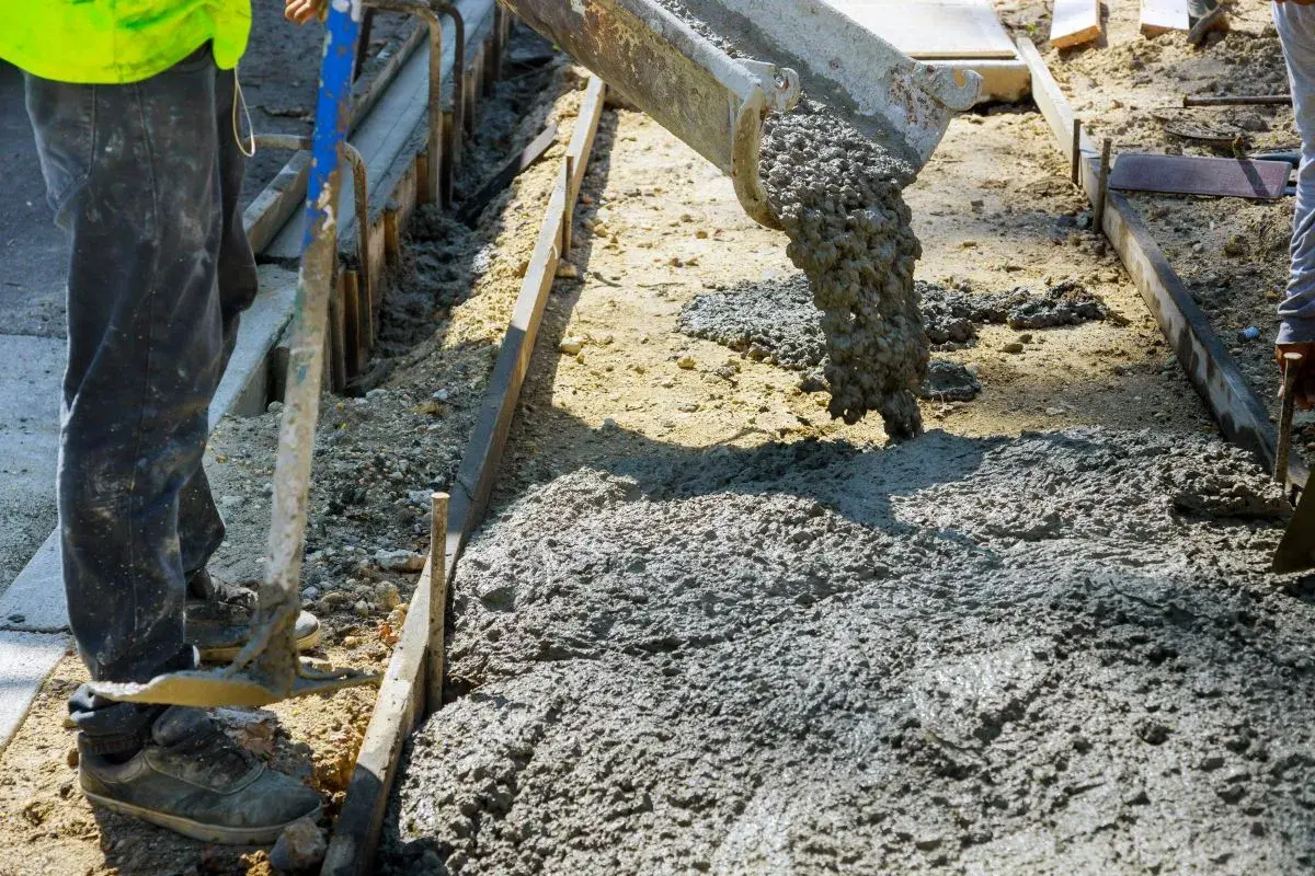 Betoniarka wylewa beton na plac budowy. Pracownik w kamizelce i spodniach przygotowuje teren. Zastanawiasz się, ile kosztuje kubik betonu?