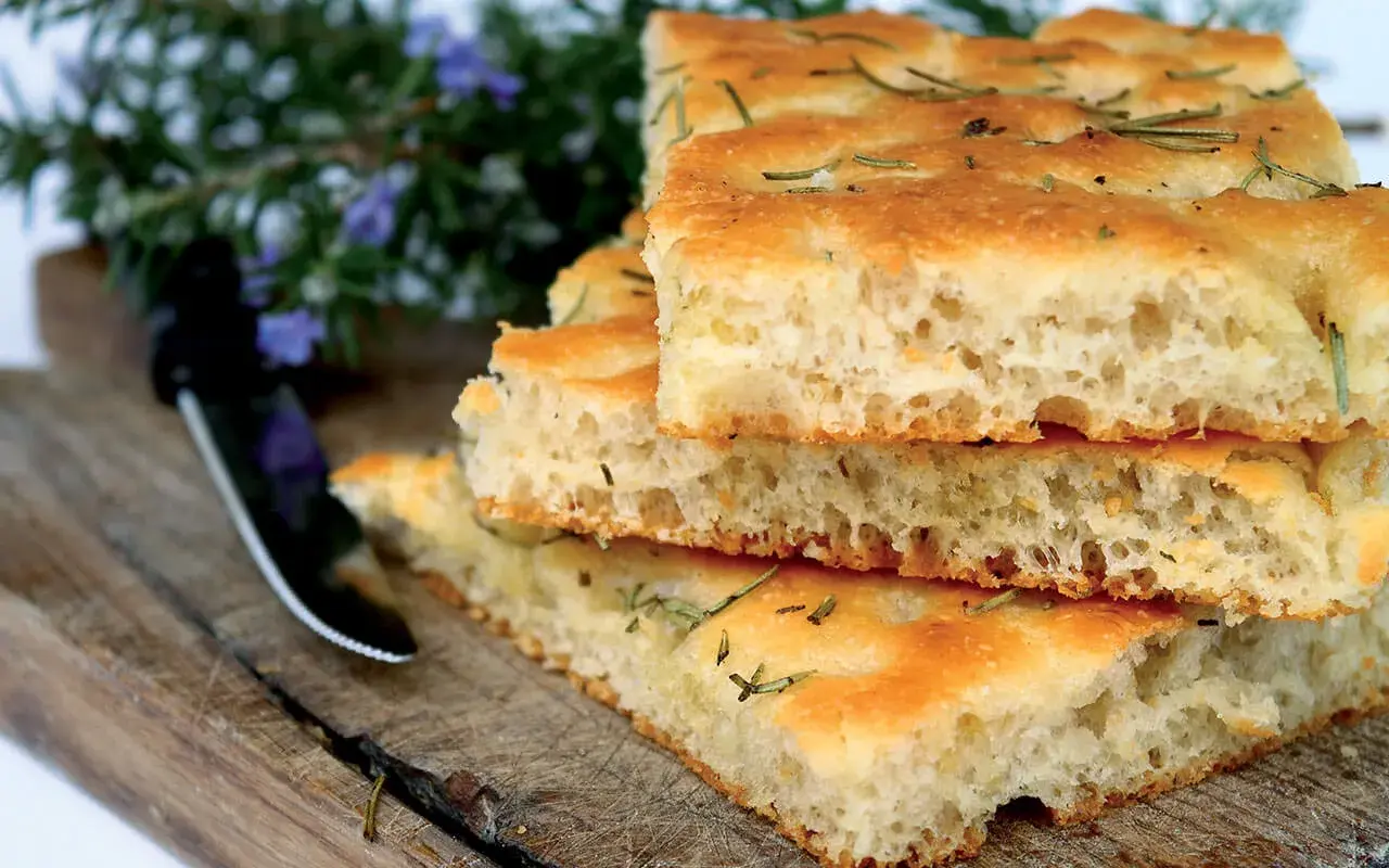 Gestapelte Focaccia-Stücke mit Rosmarin, die an das Backen von Pizza erinnern. Ein Messer liegt daneben auf einem Holzbrett.