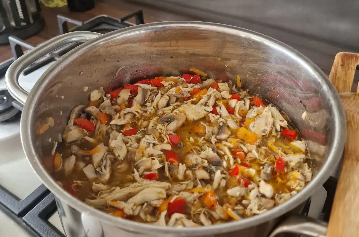 Gęsta zupa z kawałkami mięsa z rosołu, pieczarkami i kolorową papryką w metalowym garnku na kuchence.