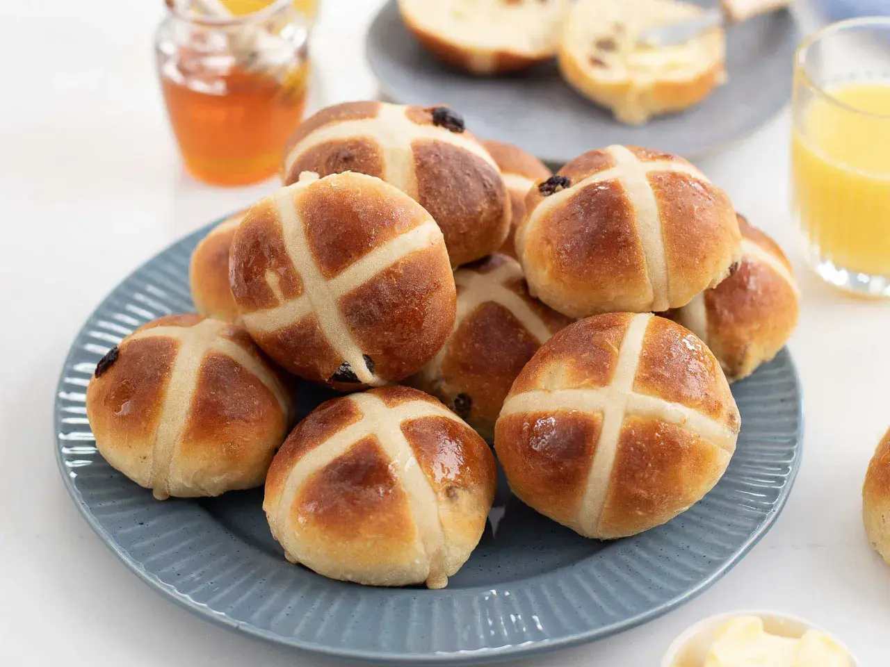 En hög med gyllene, nybakade **hot cross buns** på en blå tallrik, redo att avnjutas med honung och apelsinjuice.