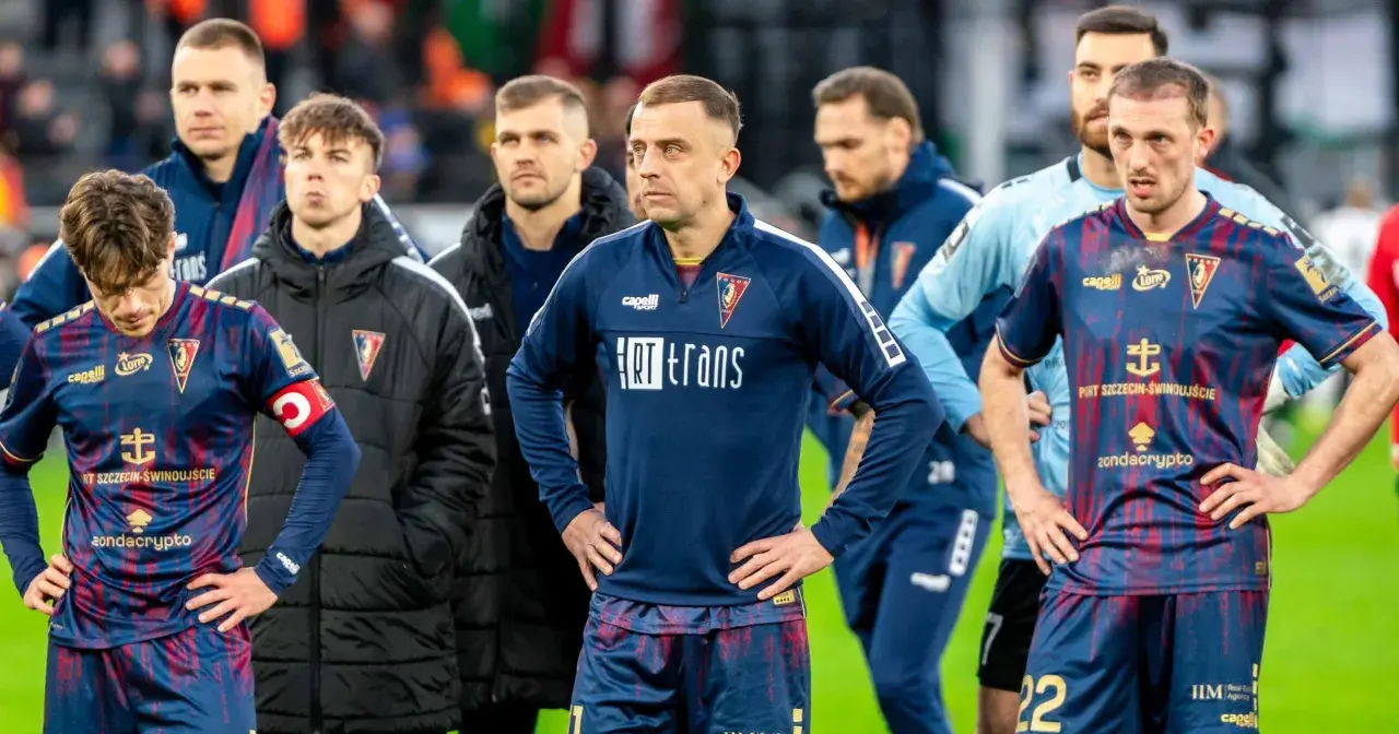 Piłkarze Pogoni Szczecin w ciemnych strojach, z widocznymi herbami klubu. W tle inni zawodnicy i sztab. Wszyscy wyglądają na zamyślonych, być może po trudnym meczu lub w oczekiwaniu na ważne pogoń szczecin - transfery.