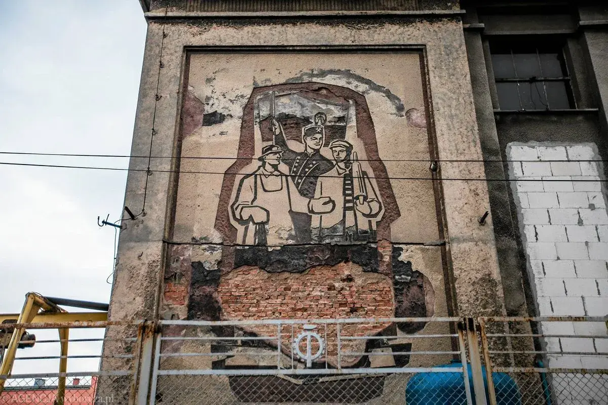 Zniszczony mural w stylu socrealizmu przedstawiający trzech robotników z karabinami.
