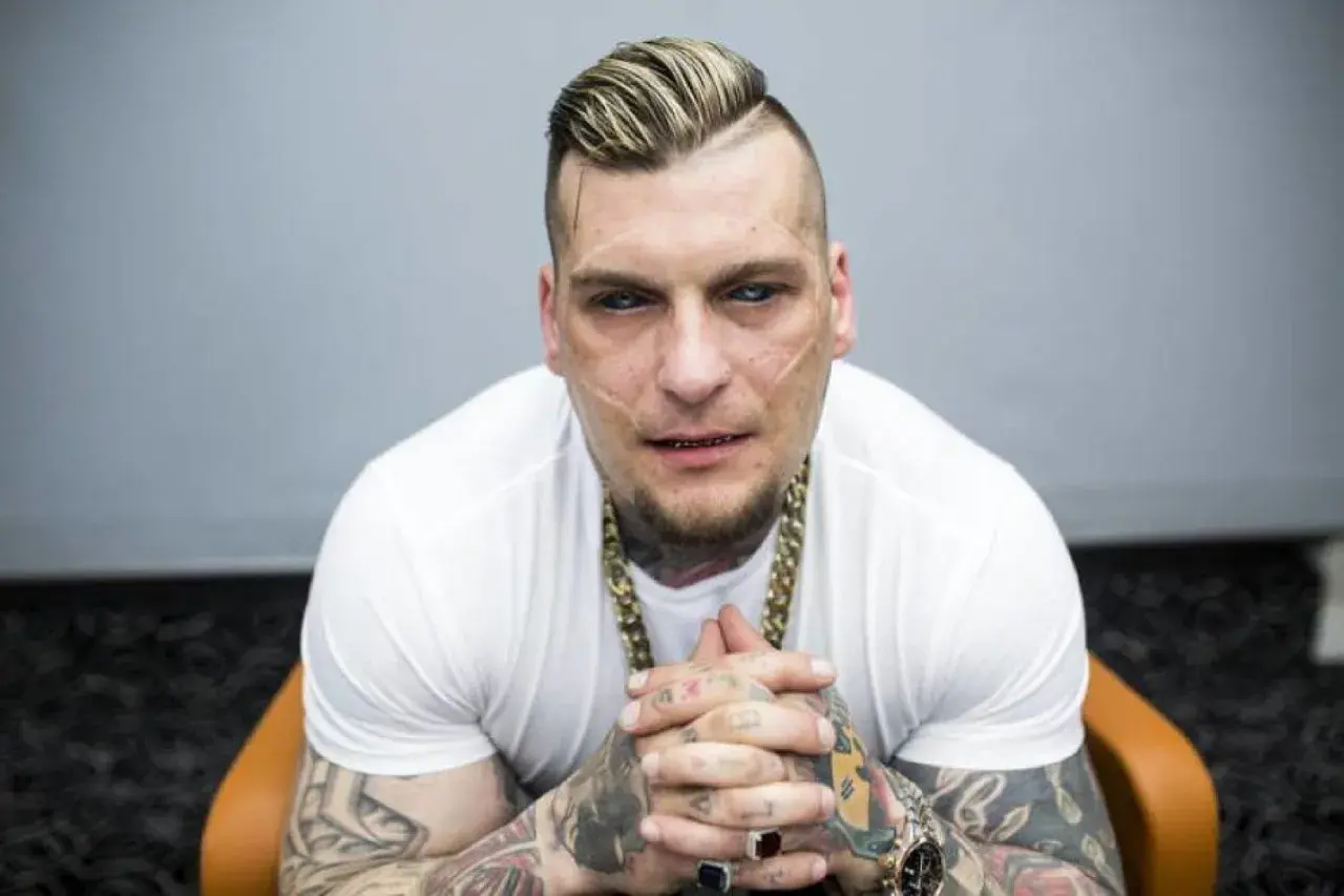 Popek kiedys, z charakterystycznymi bliznami na twarzy i tatuażami, w białej koszulce i złotym łańcuchu, siedzi na krześle.