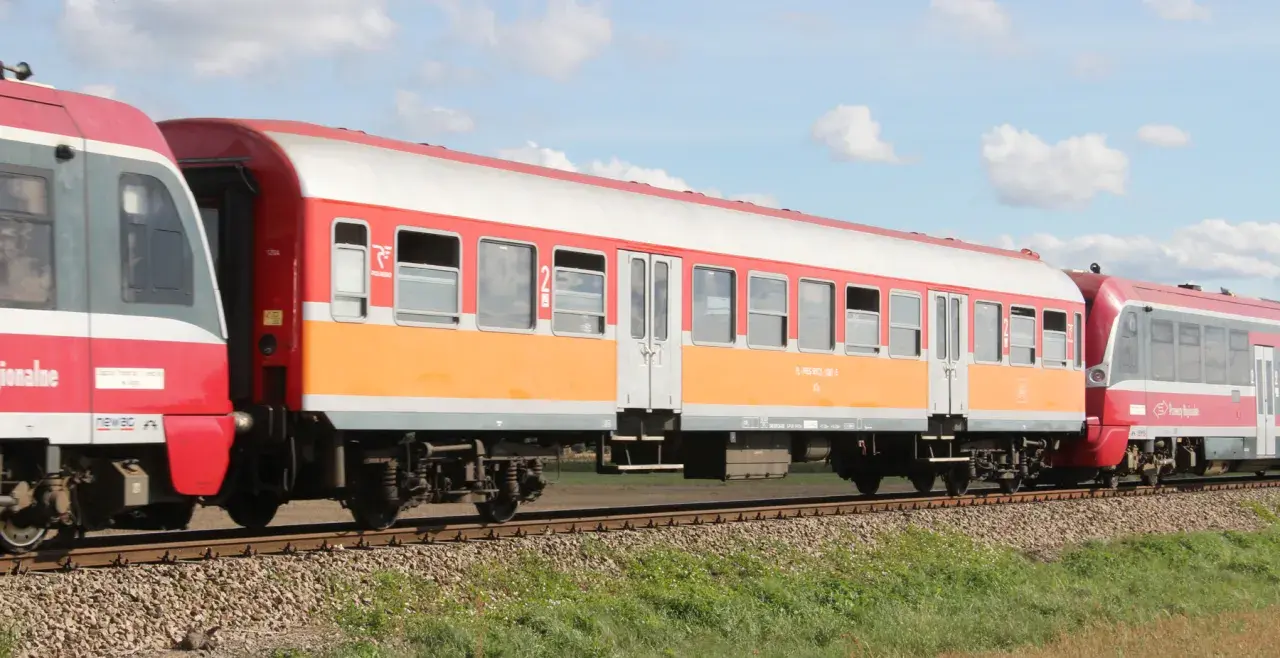 Lekki wagon pasażerski w kolorach pomarańczowo-białym i czerwonym jedzie po torach.