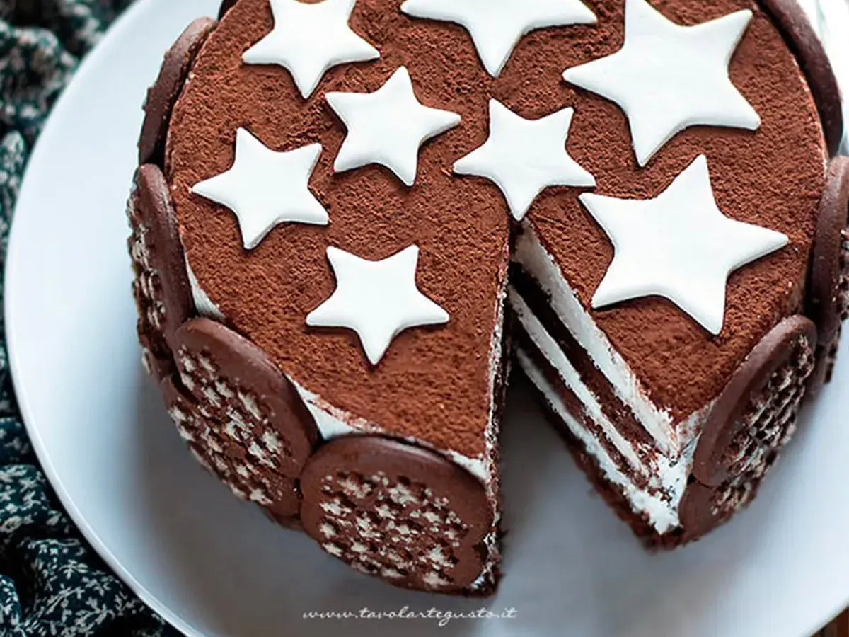 Torta Pan di Stelle ricoperta di cacao, decorata con stelle bianche e biscotti. Una fetta mostra gli strati di crema.