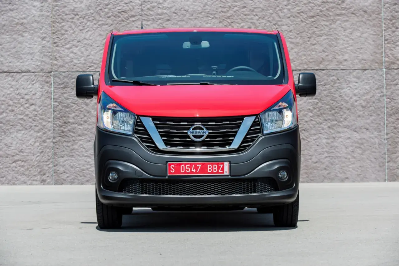 Czerwony Nissan NV300 z czarnym zderzakiem i chromowaną atrapą chłodnicy stoi przed betonową ścianą.