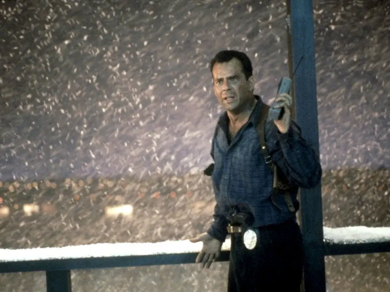John McClane, med en gammal mobiltelefon i handen, står i snöstormen. Han är en del av rollistan i Die Hard 2.