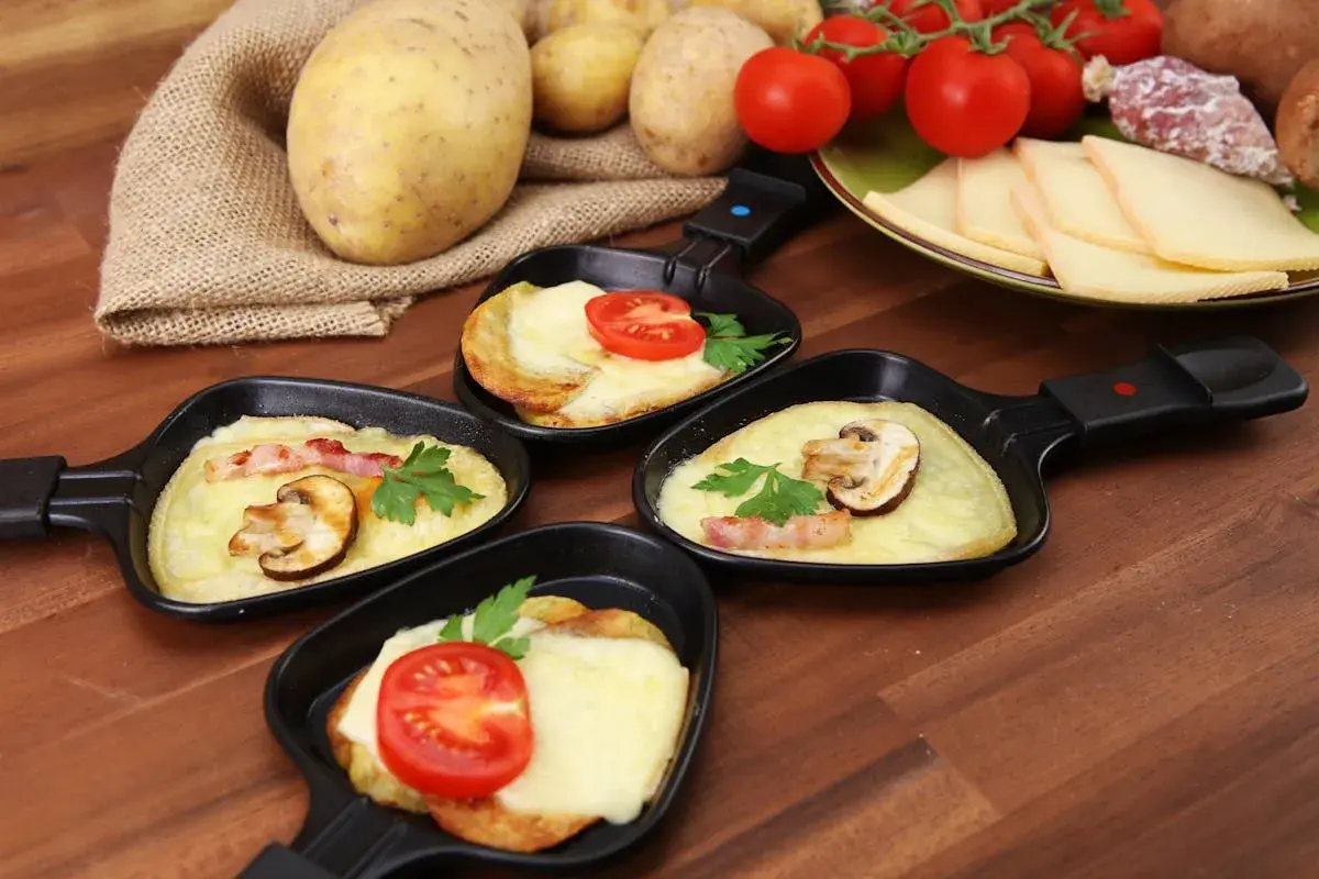 Petites poêles de raclette prêtes à être dégustées, avec pommes de terre, fromage fondu, tomates et champignons. Idéal pour savoir comment couper la raclette.