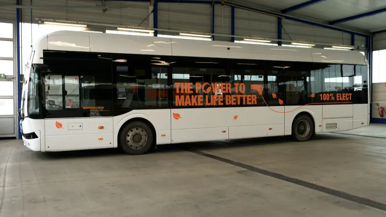 Ein weißer, moderner elektrischer Bus steht in einer Halle. Auf der Seite steht "THE POWER TO MAKE LIFE BETTER" und "100% ELECT".