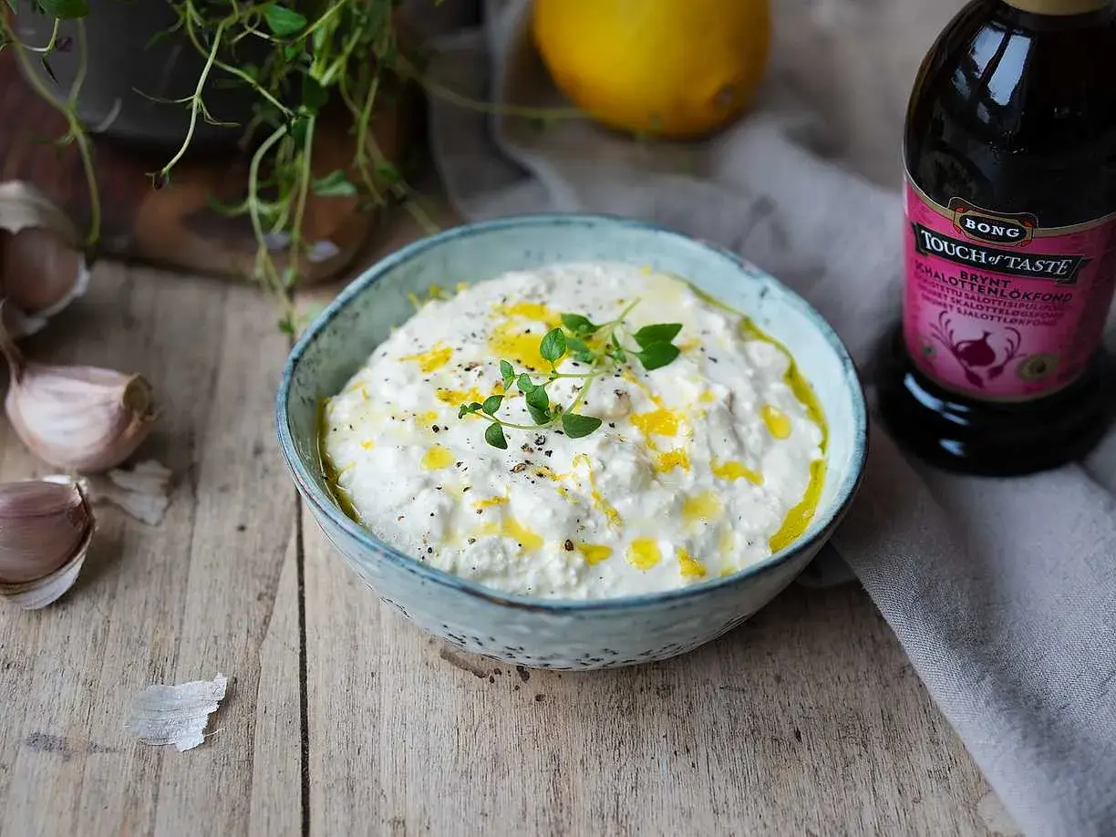 En skål med krämig, vispad fetaost toppad med olivolja och örter. Bredvid syns vitlök, citron och en flaska fond.
