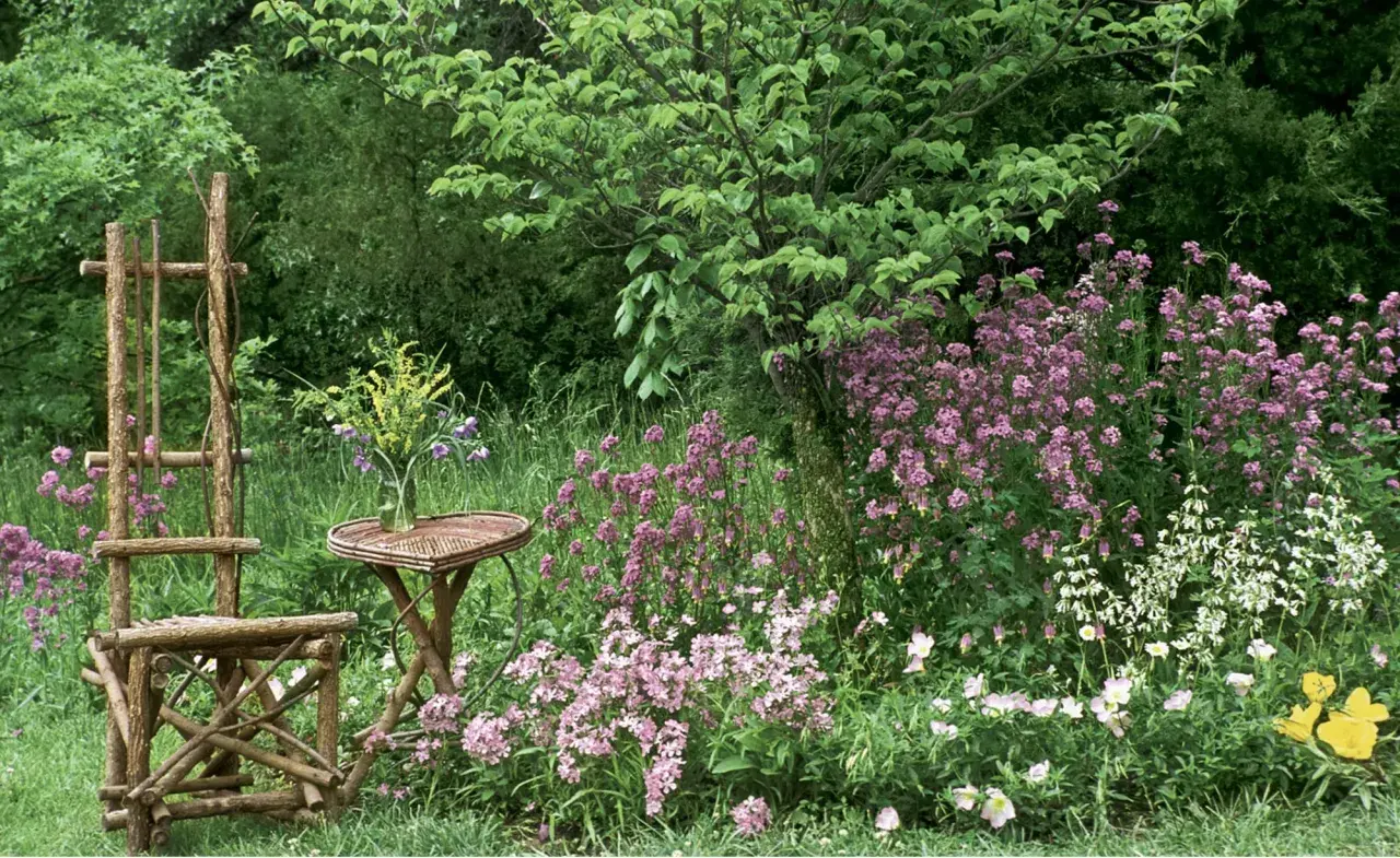 Ein rustikaler Holzstuhl und Tisch stehen vor einer üppigen, naturnahen Hecke mit rosa und weißen Blüten.