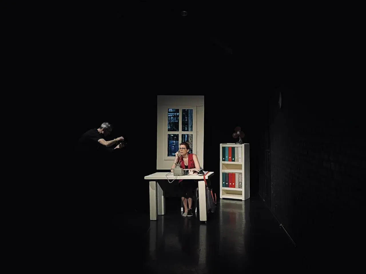 Scena w **teatrze Kana Szczecin**: kobieta przy biurku rozmawia przez telefon, mężczyzna w tle filmuje.