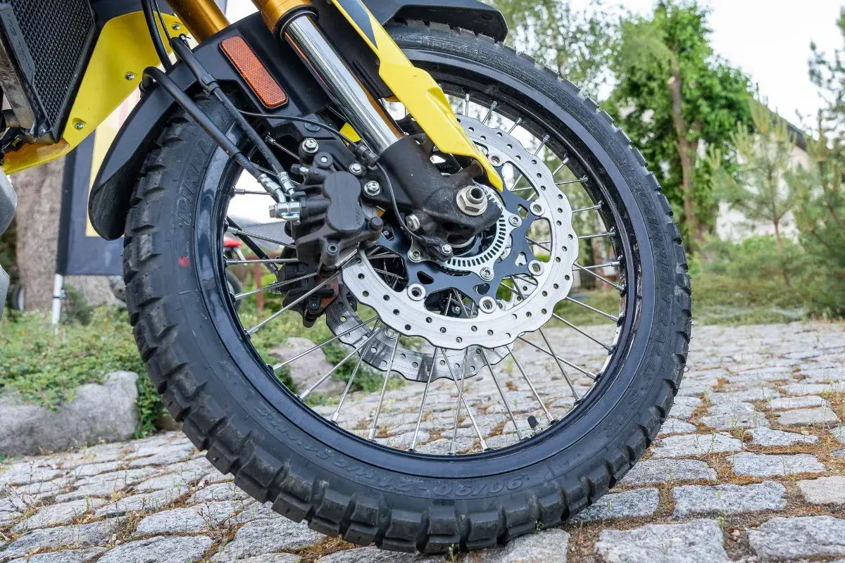 Koło motocykla z tarczą hamulcową i zaciskiem, na tle kamiennej nawierzchni i zieleni.