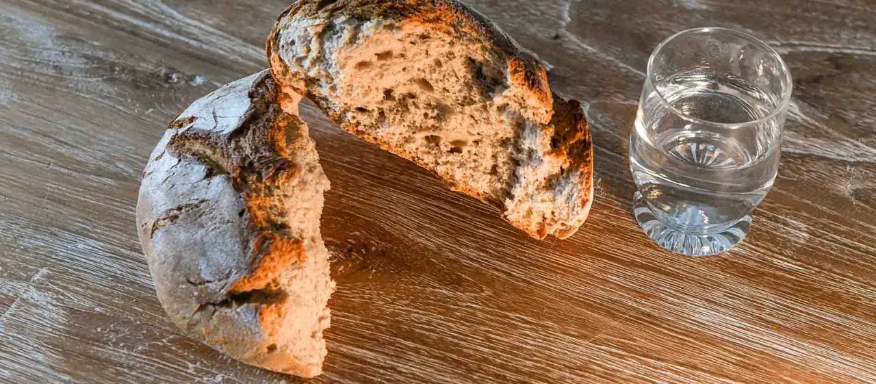 Ein aufgeschnittenes Brot, ein starkes Symbol für Nahrung und Leben, liegt neben einem Glas Wasser auf einem Holztisch.