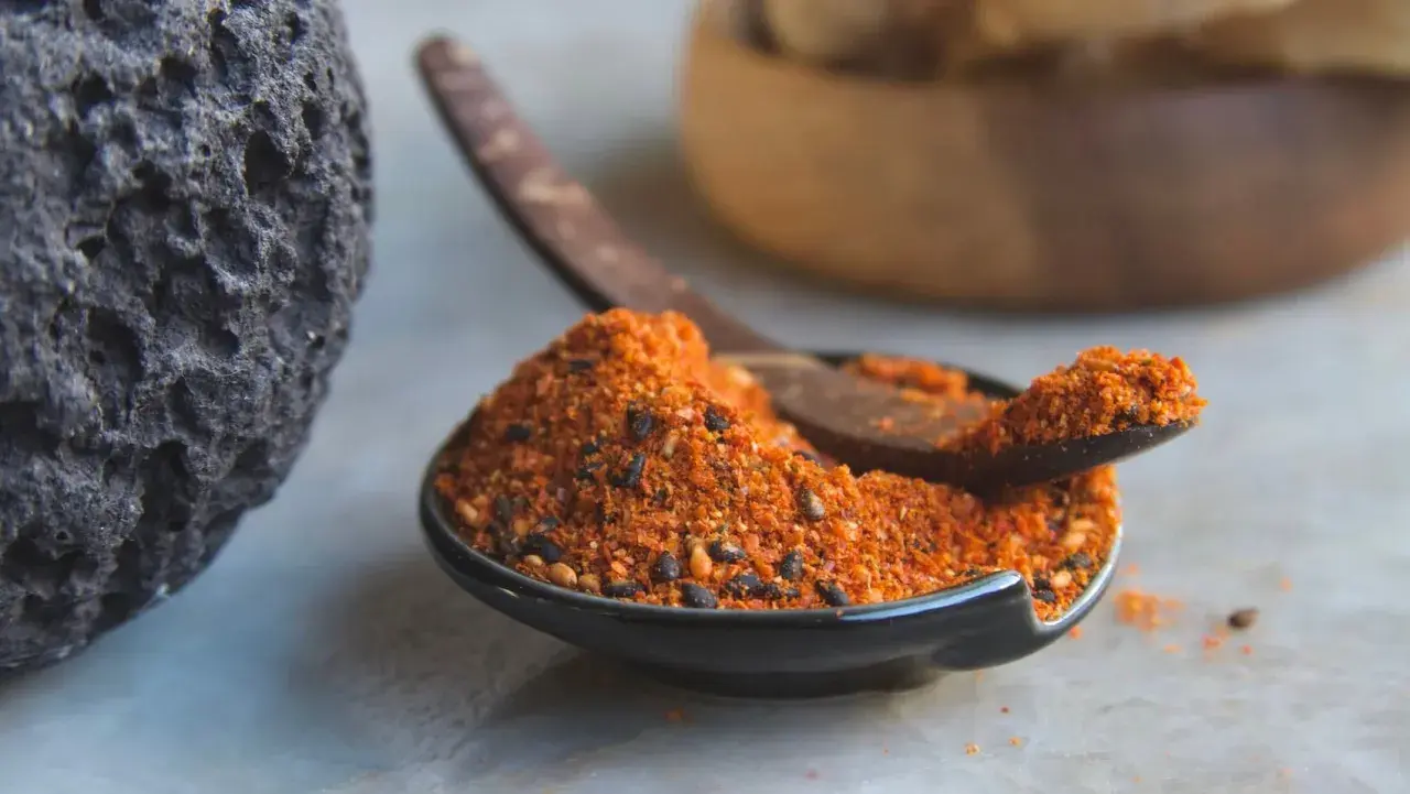 A dark, textured rock sits beside a small bowl filled with a vibrant 7 spice recipe blend. A wooden spoon rests in the bowl, ready to scoop the aromatic mixture.
