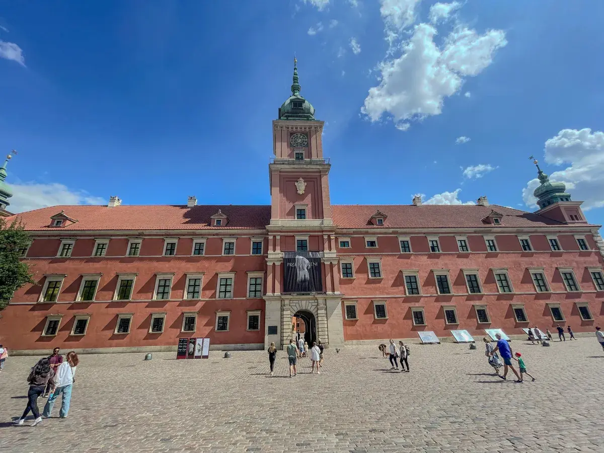 Zamek Królewski w Warszawie, z czerwoną fasadą i wieżą zegarową, na tle błękitnego nieba z chmurami. Plac przed zamkiem tętni życiem.
