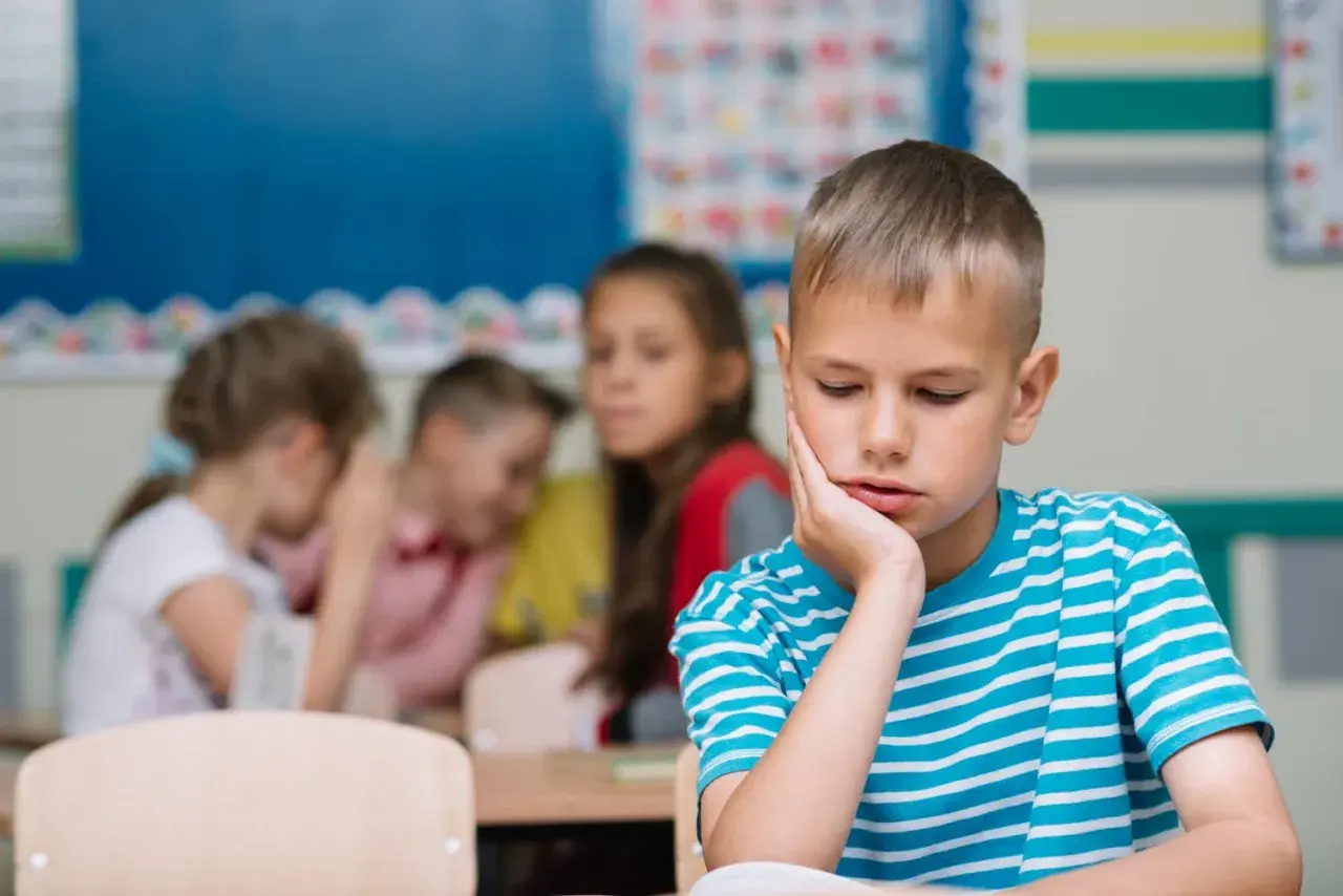Un bambino solo in classe, con la mano sul viso, mentre altri bambini ridono e parlano tra loro. Sembra che mia figlia non abbia amici.