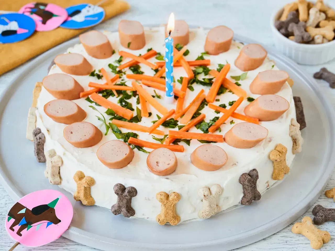 Hunde Torte Rezept einfach und schnell: Leckere Wurstscheiben, Karotten und Kräuter zieren diese festliche Hundetorte mit Kerze. Knochenförmige Kekse schmücken den Rand.