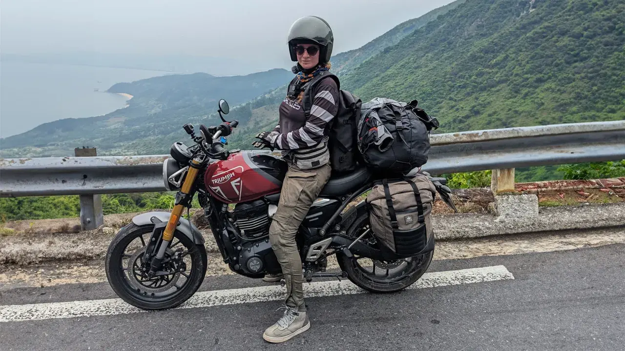 Motociclista con su equipada Triumph Tiger 400, lista para la aventura en una carretera con vistas al mar y montañas.