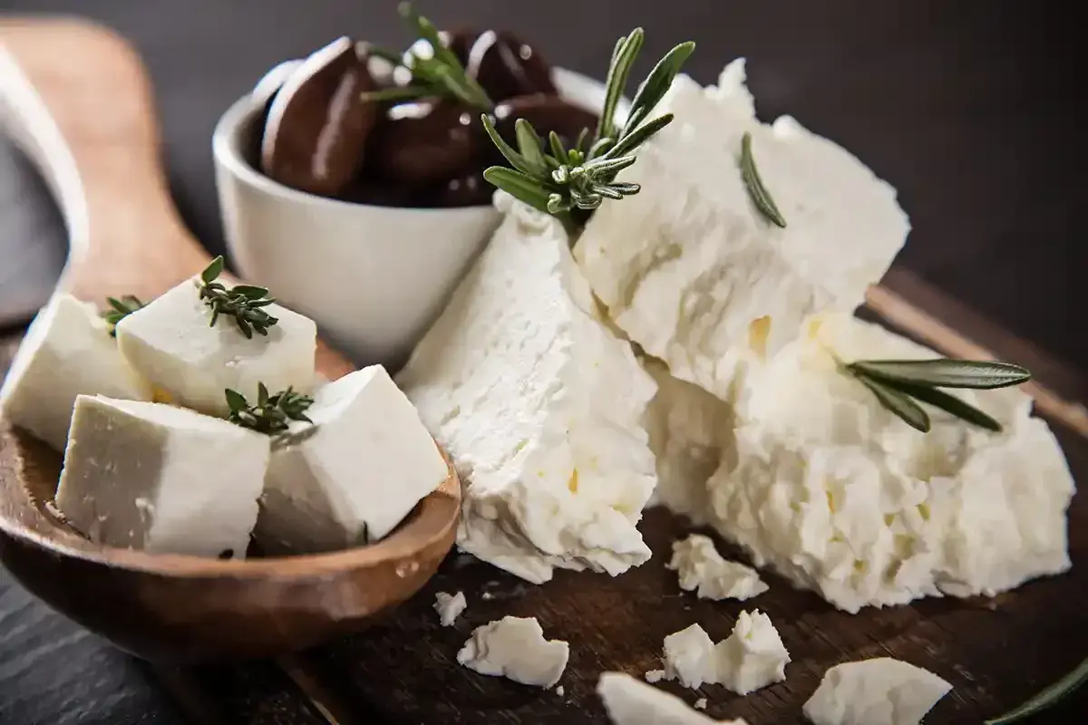Feta, un formaggio ricco di proteine per 100g, servito con olive nere e rametti di rosmarino su un tagliere di legno.