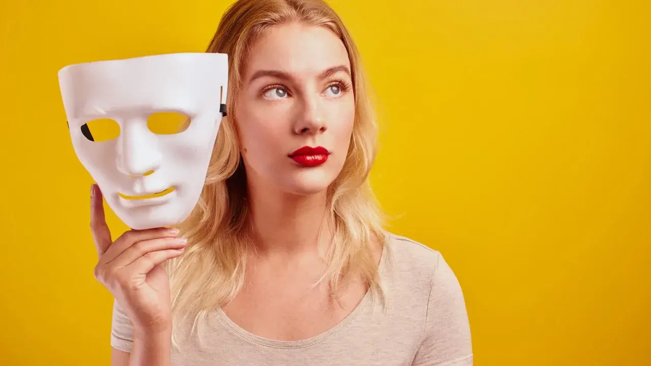Blondynka z czerwoną szminką trzyma białą maskę, jakby zastanawiała się, czy być ekstrawertykiem, czy ukryć swoją prawdziwą twarz.