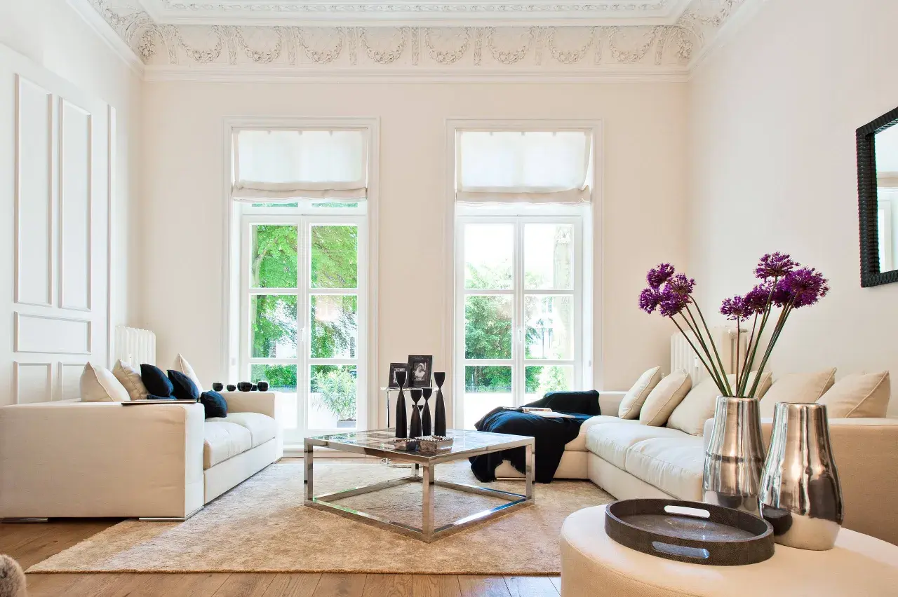 Helles Wohnzimmer im Hamptons-Stil mit beigen Sofas, einem Chrom-Couchtisch und großen Fenstern, die ins Grüne blicken.