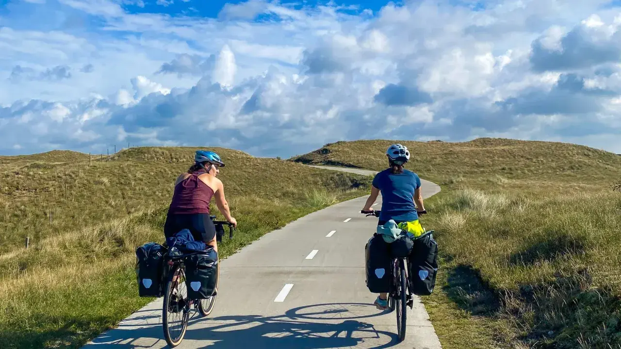 Two cyclists with panniers enjoy a scenic path through grassy dunes, perfect for Netherlands bike trips.
