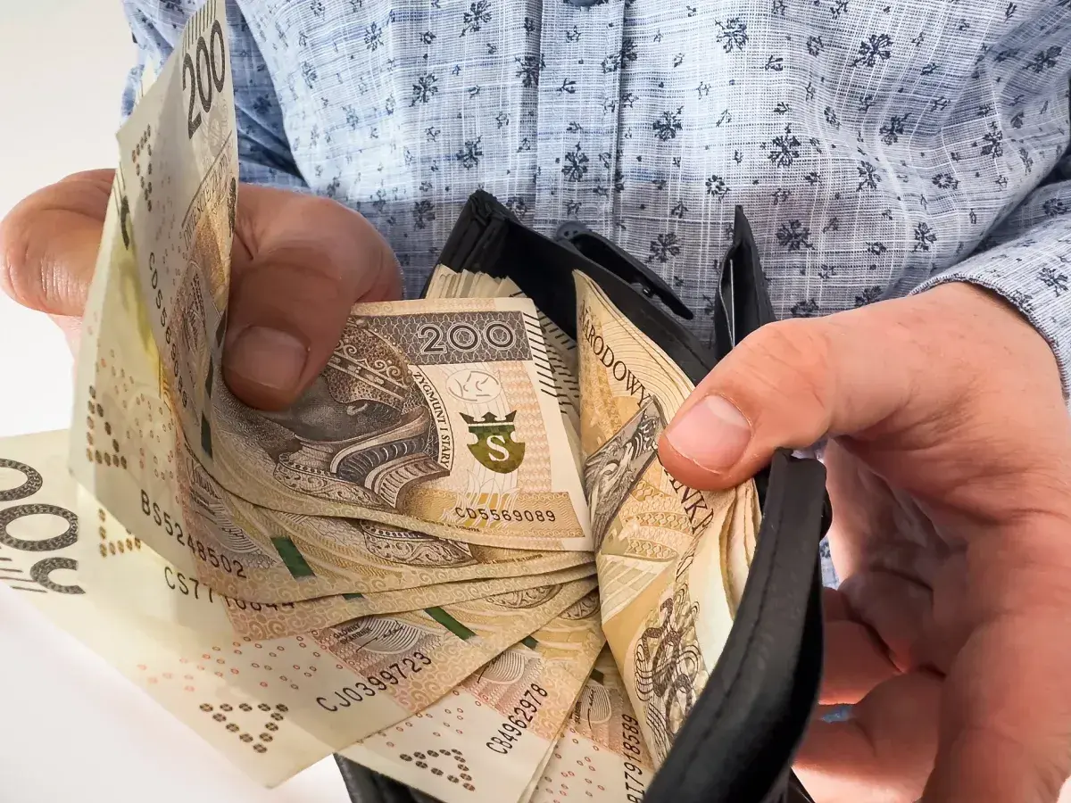Ręce trzymają portfel z banknotami 200 zł. Ile trzeba przepracować, by tyle zarobić?