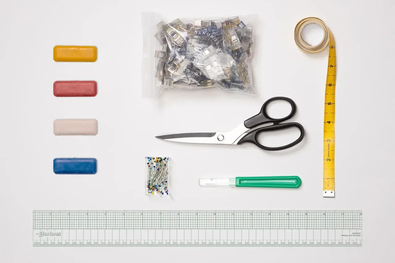 A collection of essential sewing tools, including colorful clips, pins, scissors, a measuring tape, a seam ripper, and a ruler, are laid out on a white surface.