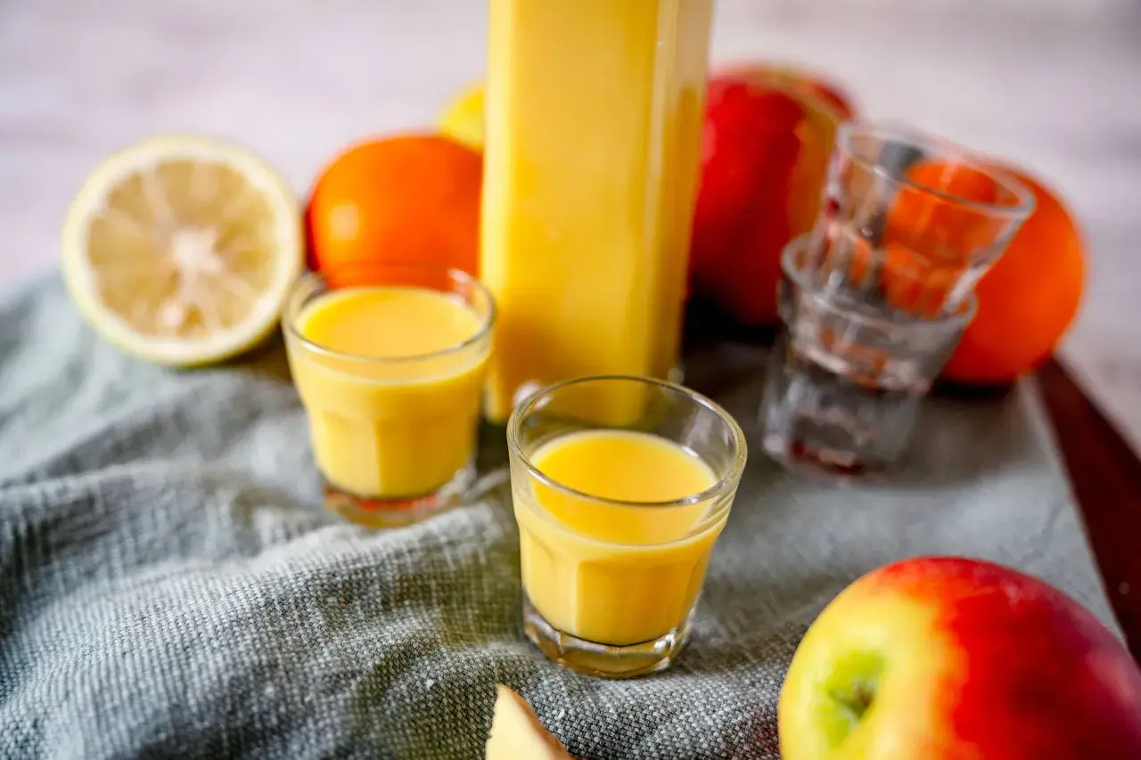 Frische Zutaten für Ingwer Shots selber machen: Zitrone, Orangen, Äpfel und ein Glas voller goldgelbem Shot.