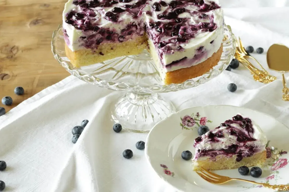 Ein köstliches Heidelbeertorte Rezept: saftiger Boden, cremige Füllung und frische Heidelbeeren. Ein Stück auf einem Teller mit goldenem Besteck.