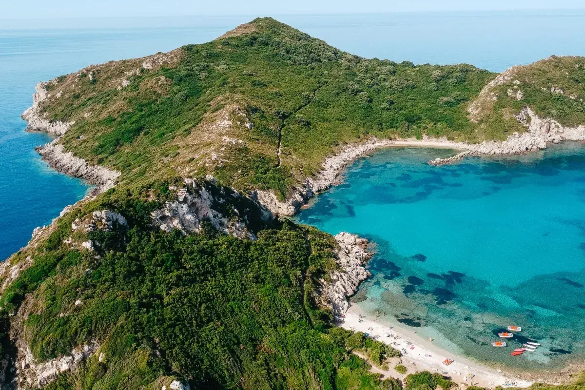 Idealna pogoda na Korfu we wrześniu: turkusowa zatoka, zielone wzgórza i piaszczysta plaża z łódkami.