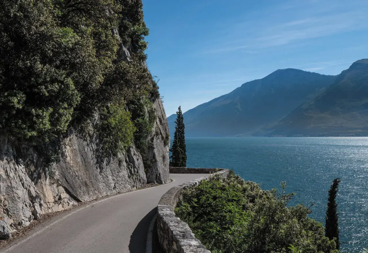 Strada panoramica lungo il lago, con rocce a sinistra e montagne sullo sfondo. La gardesana occidentale offre scorci mozzafiato.