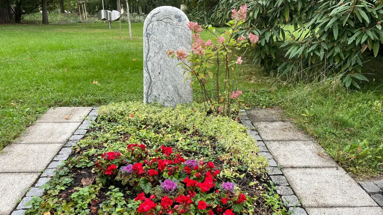Ein Grabstein mit Baumgravur, umgeben von bunten Blumen und immergrünen Pflanzen. Dauerhafte Grabbepflanzung Beispiele für eine liebevolle Gestaltung.