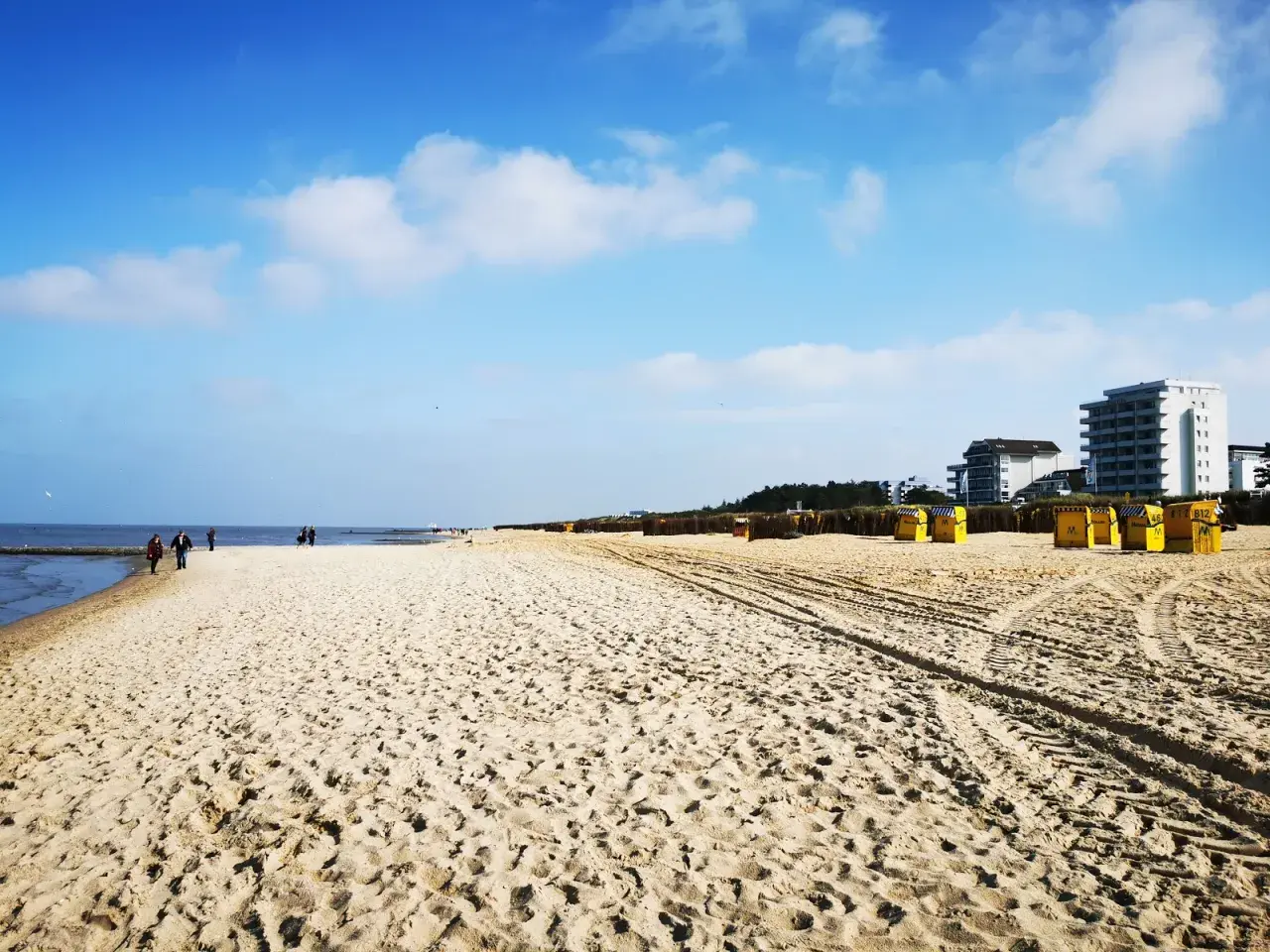 Ein weiter, sonniger Duhnen Strand mit gelben Strandkörben und einigen Spaziergängern. Im Hintergrund moderne Gebäude.