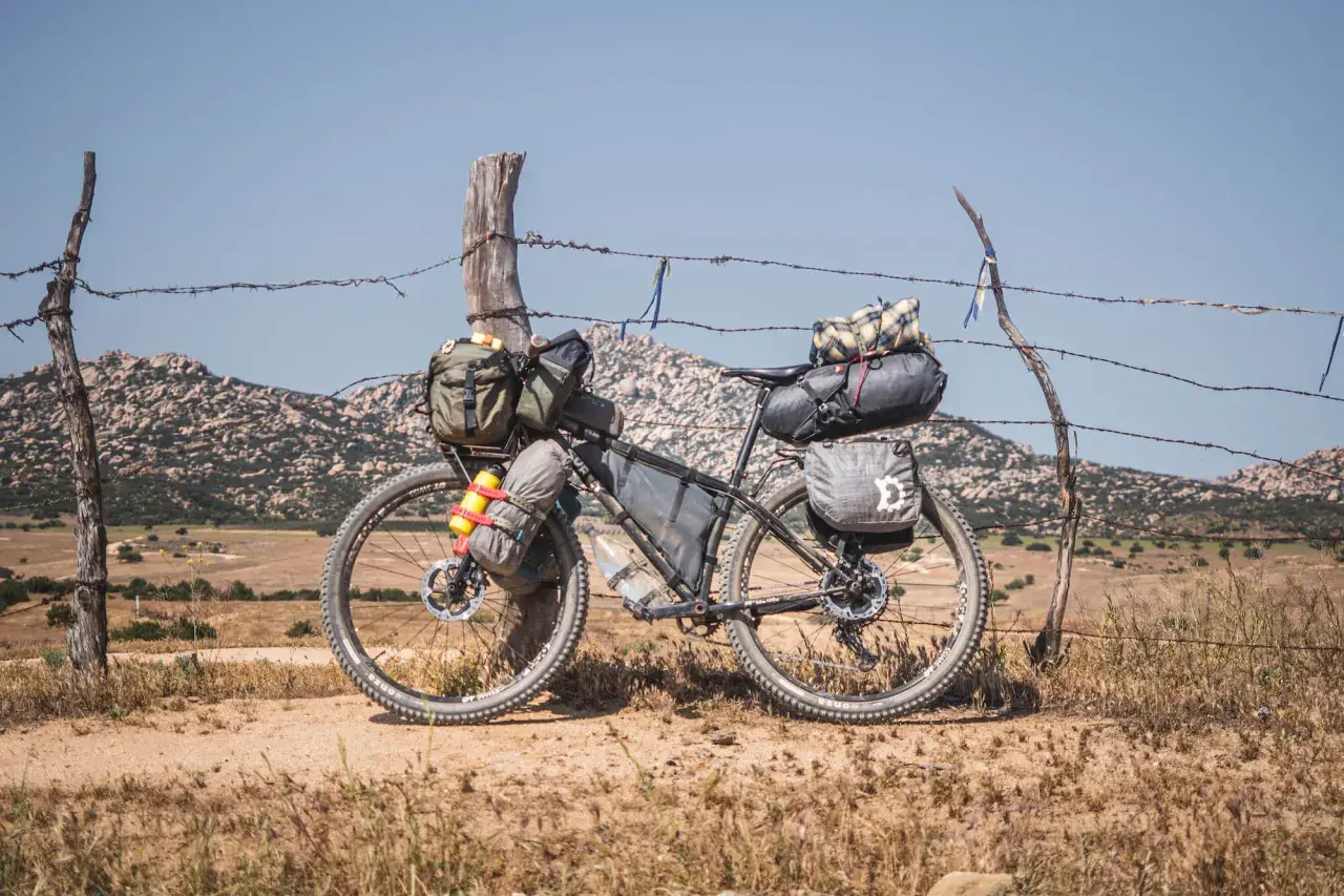 Bicicleta lista para bike packing, cargada con alforjas y bolsas, apoyada en un poste de alambre de espino, con paisaje montañoso al fondo.