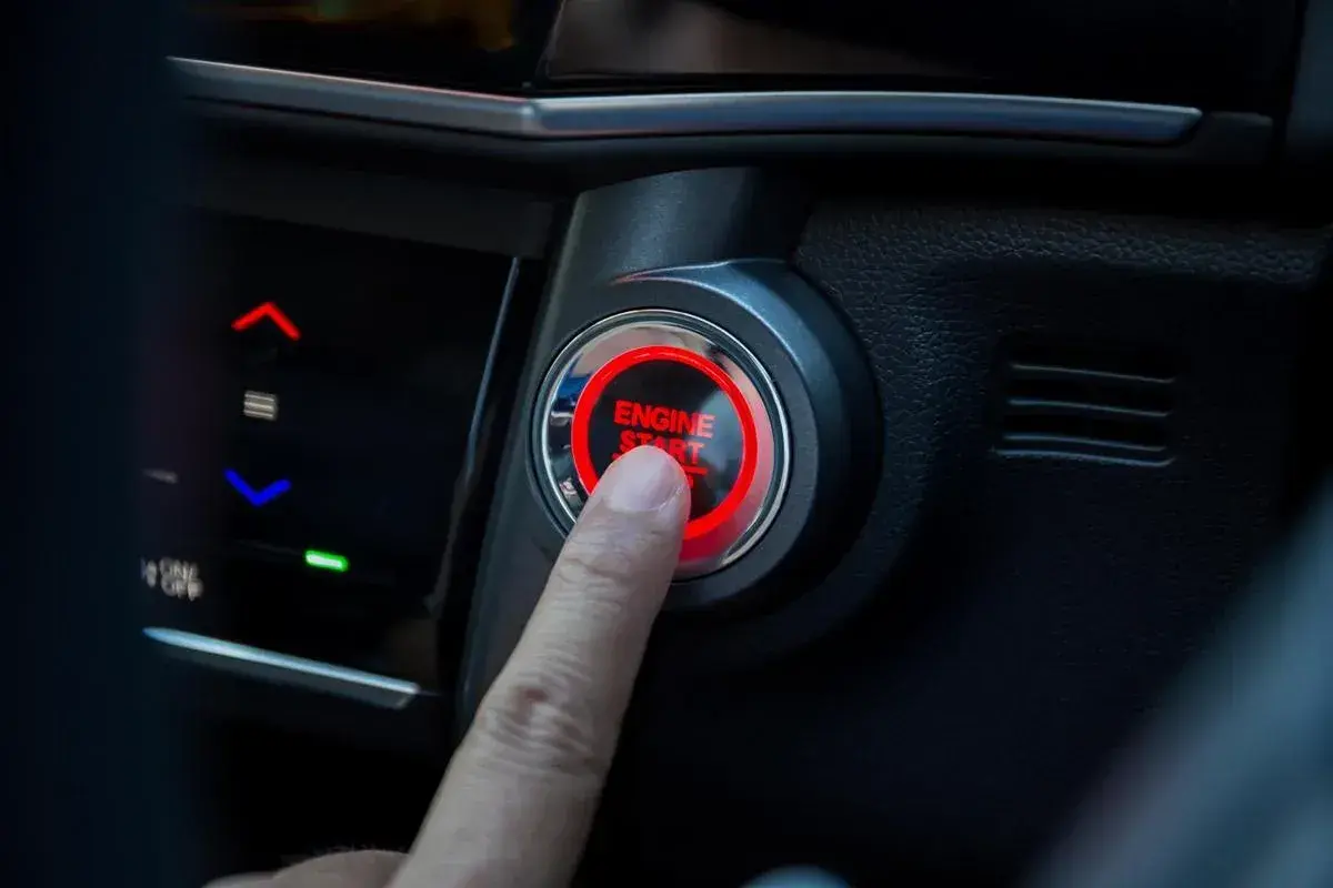 Un doigt appuie sur le bouton "ENGINE START" qui clignote en rouge. La voiture est prête à démarrer.