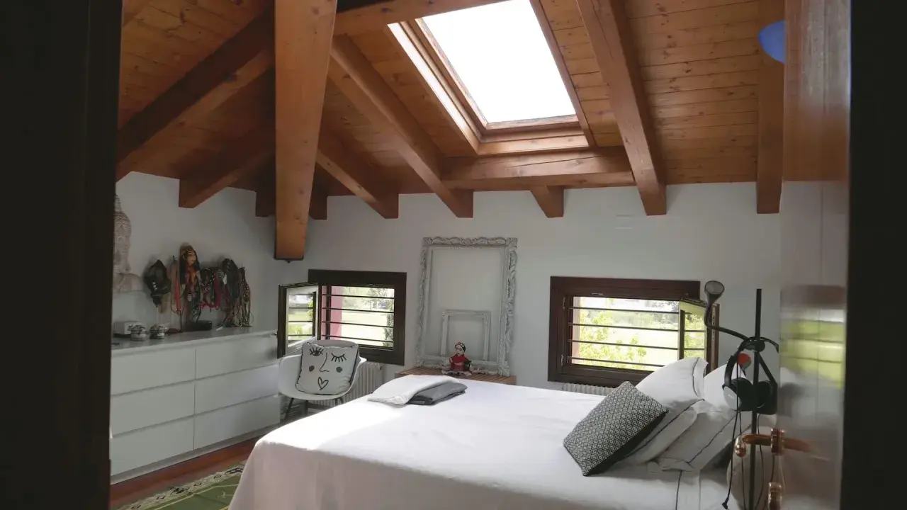 Camera da letto mansardata con travi a vista in legno. Idee colori per una mansarda luminosa e accogliente, con arredi bianchi e dettagli etnici.