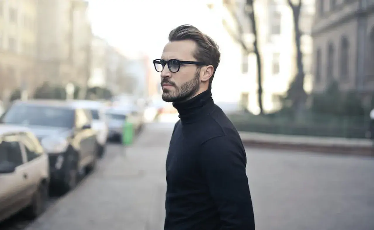 Elegancki mężczyzna w okularach i czarnym golfie, zamyślony, jakby planował zagrywki zakochanego faceta.