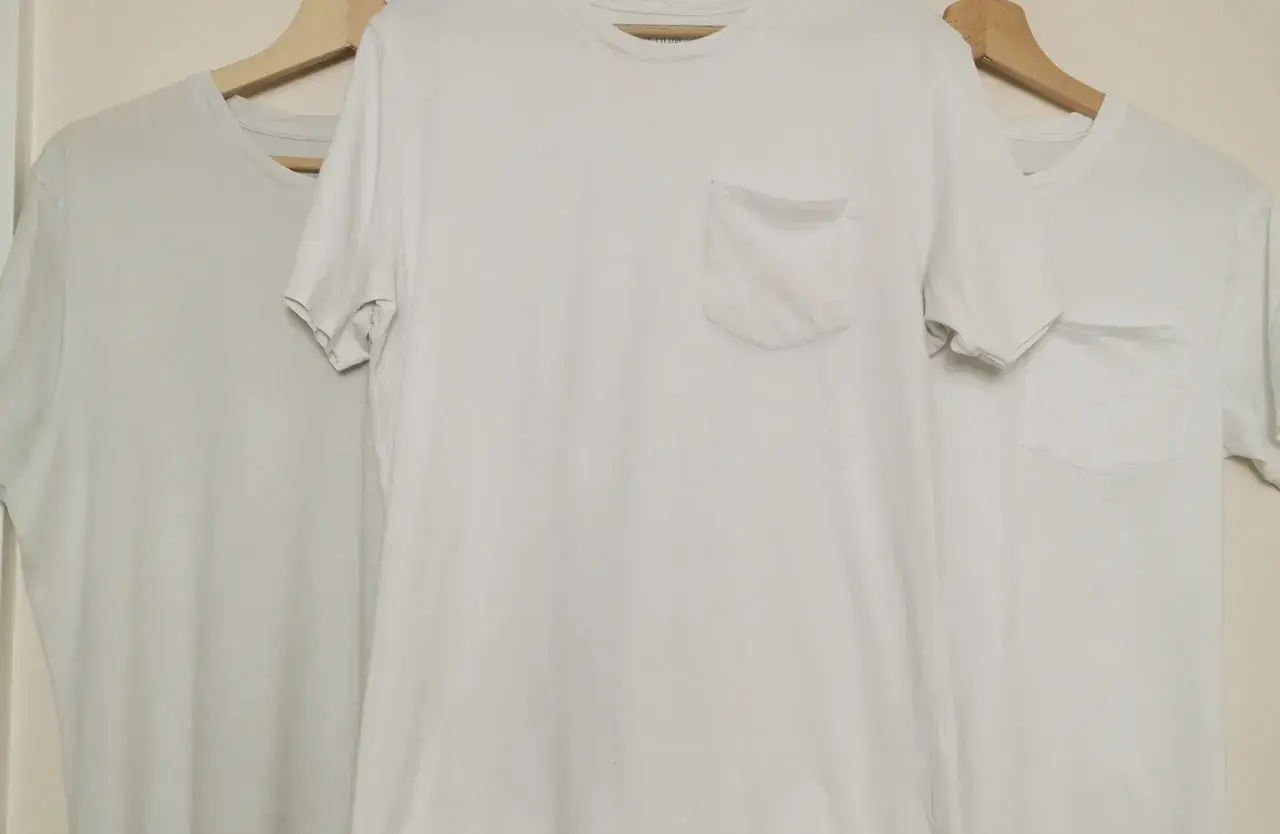 Trois t-shirts blancs sur cintres en bois. Le t-shirt du milieu a une poche poitrine. Idéal pour apprendre comment blanchir un pull blanc.