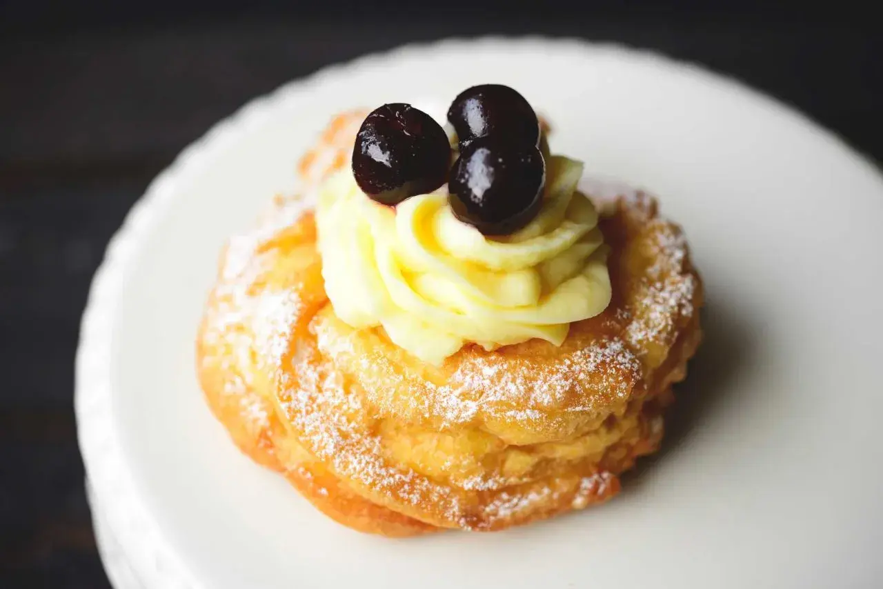 Zeppole fritte gonfie, soffice pasta con crema pasticcera e ciliegie candite, spolverate di zucchero a velo.