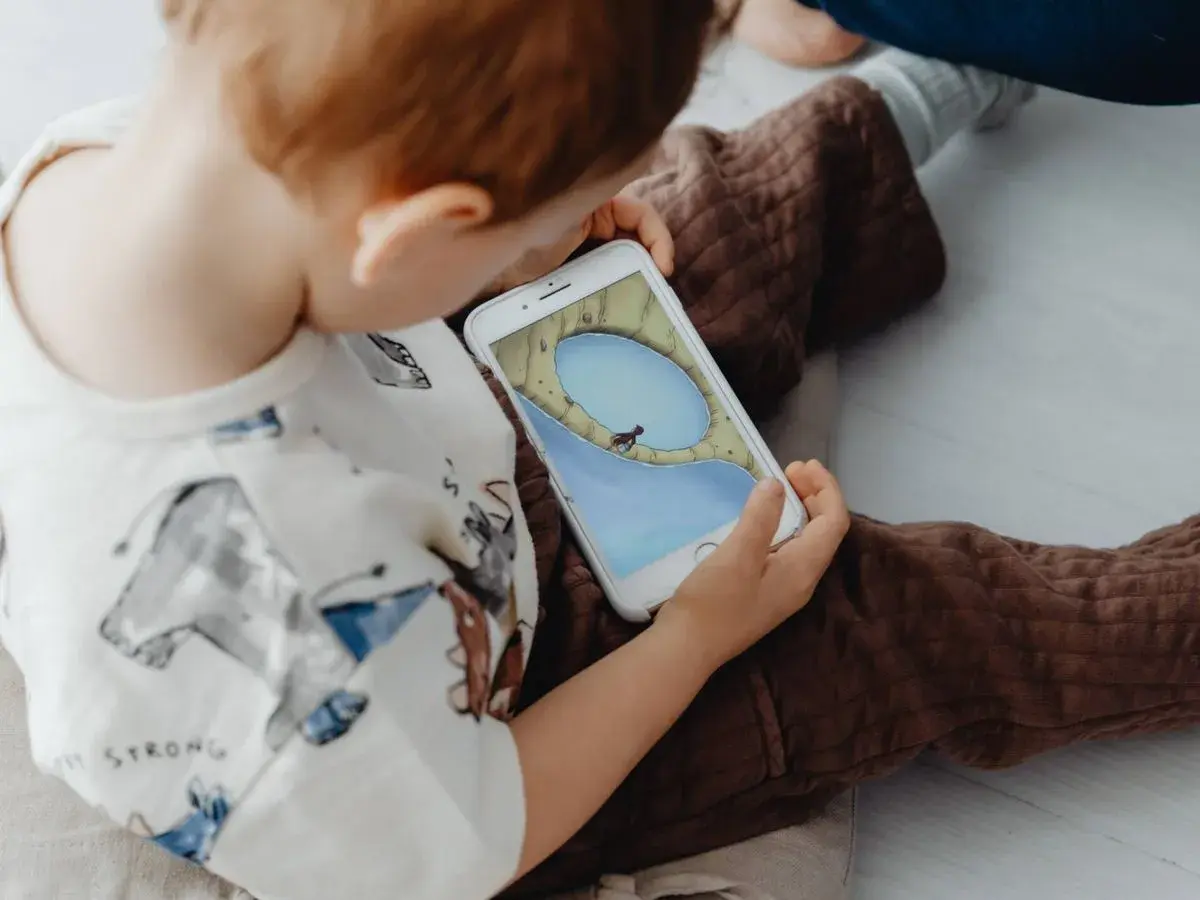 Un niño, absorto en su móvil, parece un niño adicto al móvil. La pantalla muestra un juego con un personaje en un paisaje acuático.