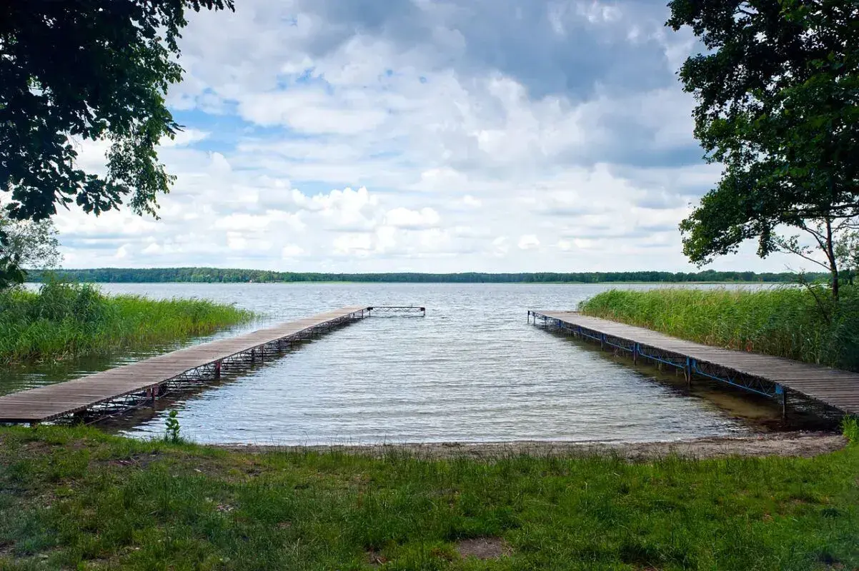 Dwa drewniane pomosty prowadzące na spokojne jezioro, otoczone zielenią. Idealne miejsce na relaks i podziwianie przyrody, jedna z wielu atrakcji w Boszkowie.
