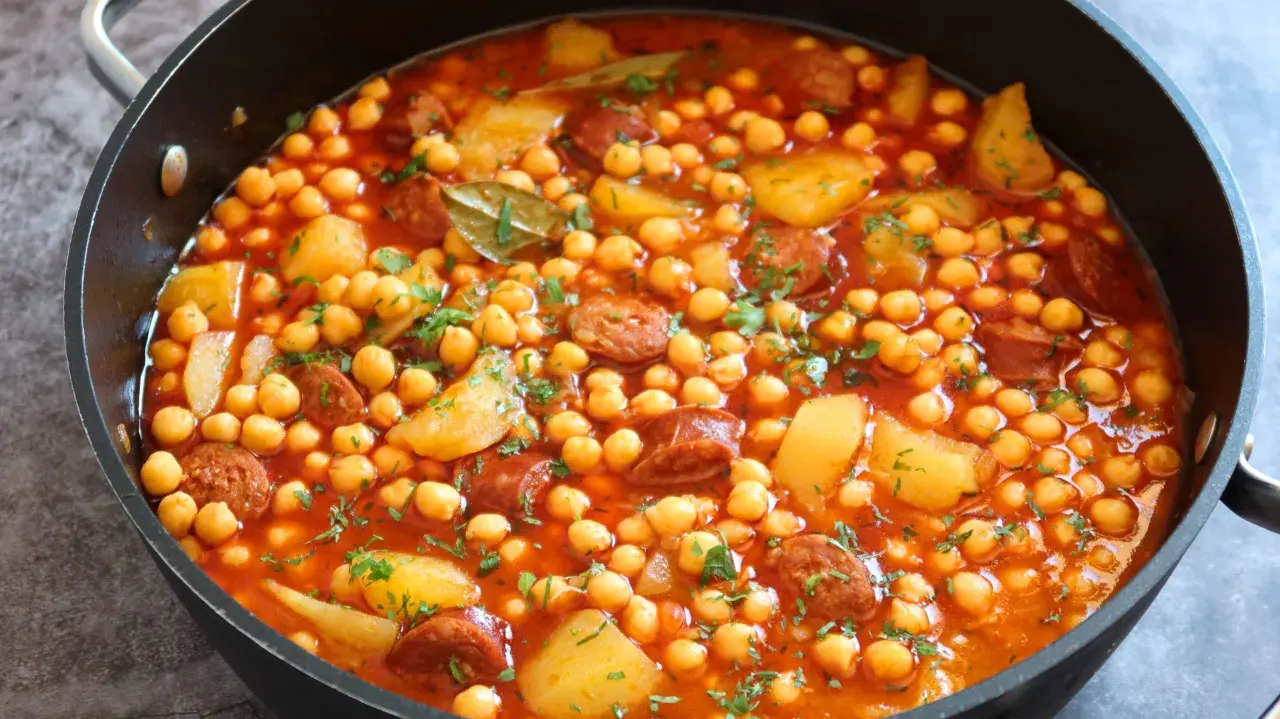 Un plato humeante de garbanzos con chorizo arguiñano, patatas y perejil picado en una sartén negra.
