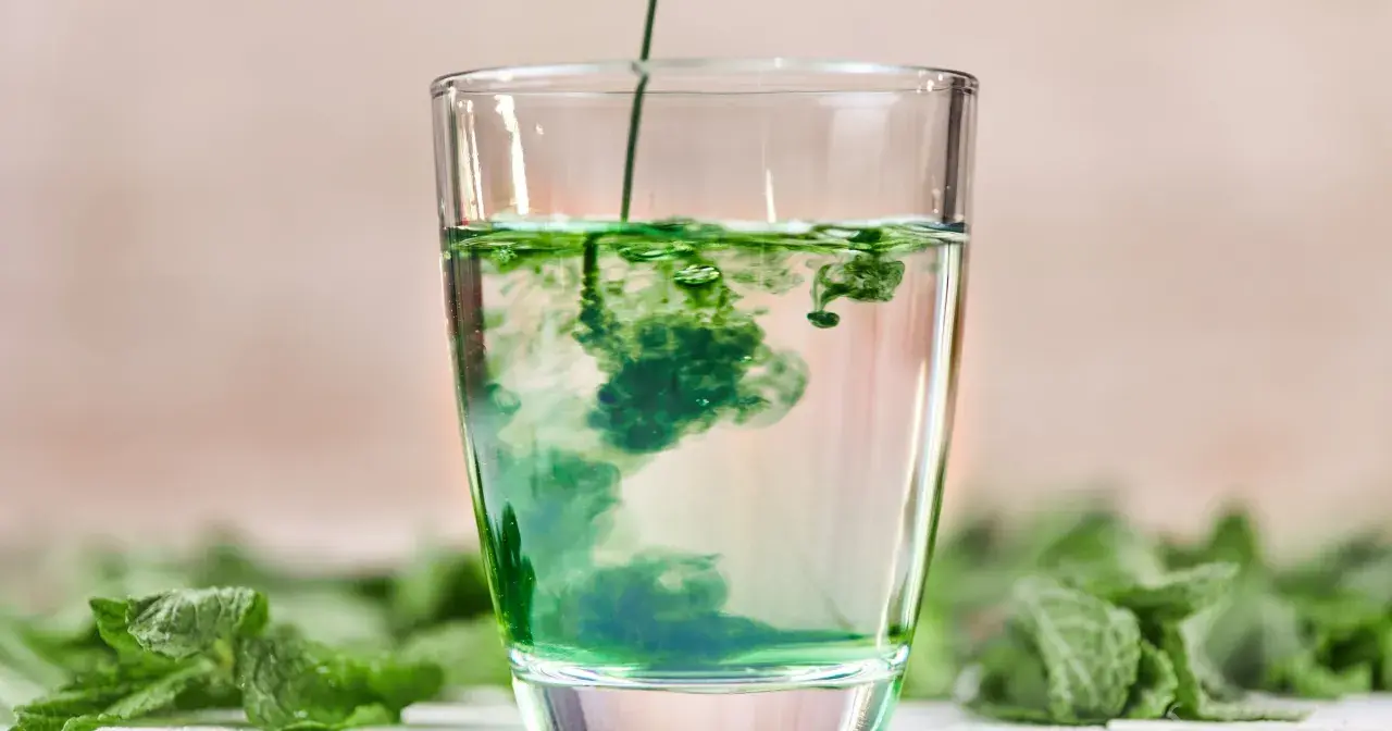 Zielony płyn rozpuszcza się w szklance wody, tworząc wiry. To naturalny chlorofil do picia, otoczony świeżymi liśćmi mięty.