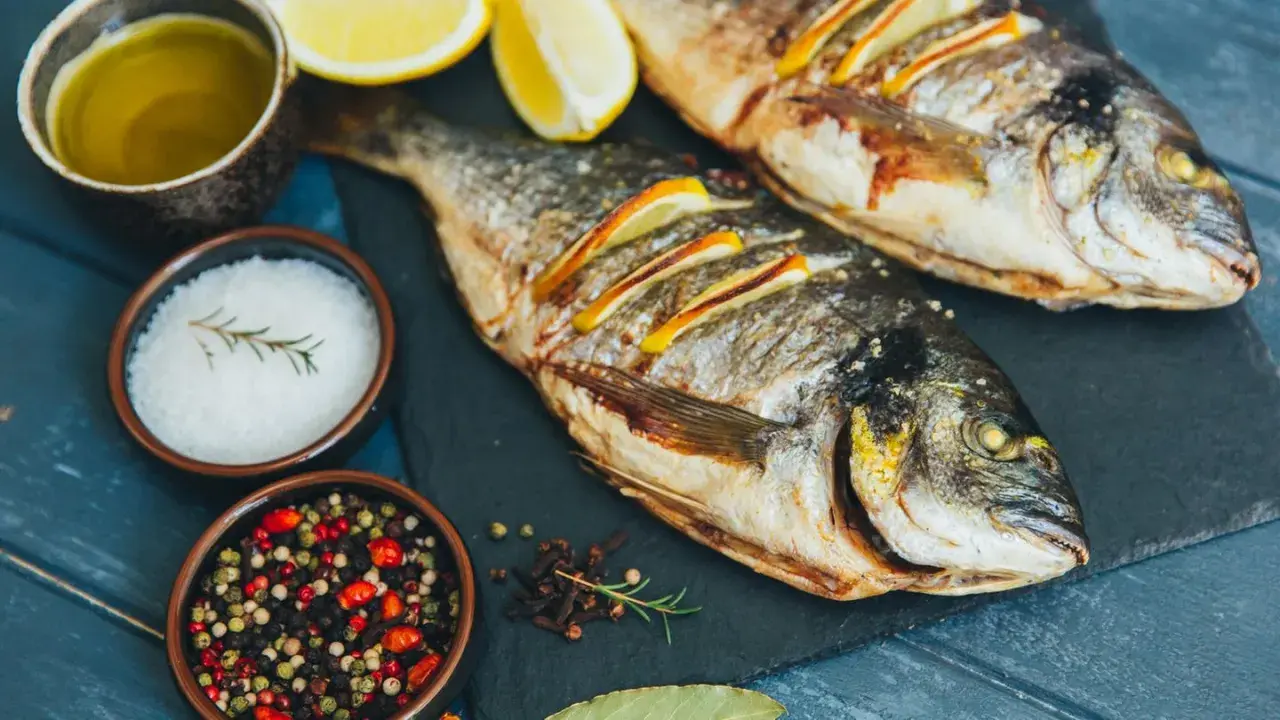 Zwei gebratene Dorade Fische mit Zitronenscheiben, Olivenöl, Salz und Pfefferkörnern auf einem dunklen Brett.